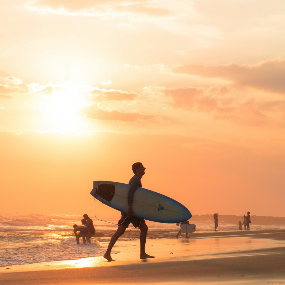 A surfer walking out of golden water with a board under one arm at sunset